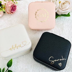 K&ouml;nnte beinhalten: Drei kleine quadratische Schmuckk&auml;stchen mit unterschiedlichfarbigen Deckeln. Die K&auml;stchen sind mit Namen und Initialen personalisiert. Das K&auml;stchen links ist wei&szlig; mit dem Namen "abbie" in goldenen Buchstaben. Das K&auml;stchen in der Mitte ist rosa mit den Initialen "GM" in einem goldenen Kranz. Das K&auml;stchen rechts ist schwarz mit dem Namen "Sarah" in goldenen Buchstaben.
