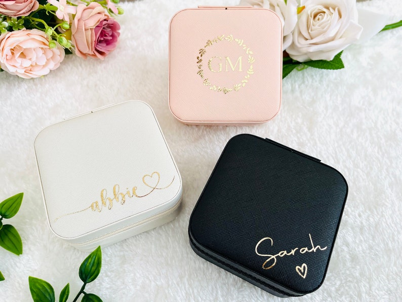 K&ouml;nnte beinhalten: Drei kleine quadratische Schmuckk&auml;stchen mit unterschiedlichfarbigen Deckeln. Die K&auml;stchen sind mit Namen und Initialen personalisiert. Das K&auml;stchen links ist wei&szlig; mit dem Namen "abbie" in goldenen Buchstaben. Das K&auml;stchen in der Mitte ist rosa mit den Initialen "GM" in einem goldenen Kranz. Das K&auml;stchen rechts ist schwarz mit dem Namen "Sarah" in goldenen Buchstaben.