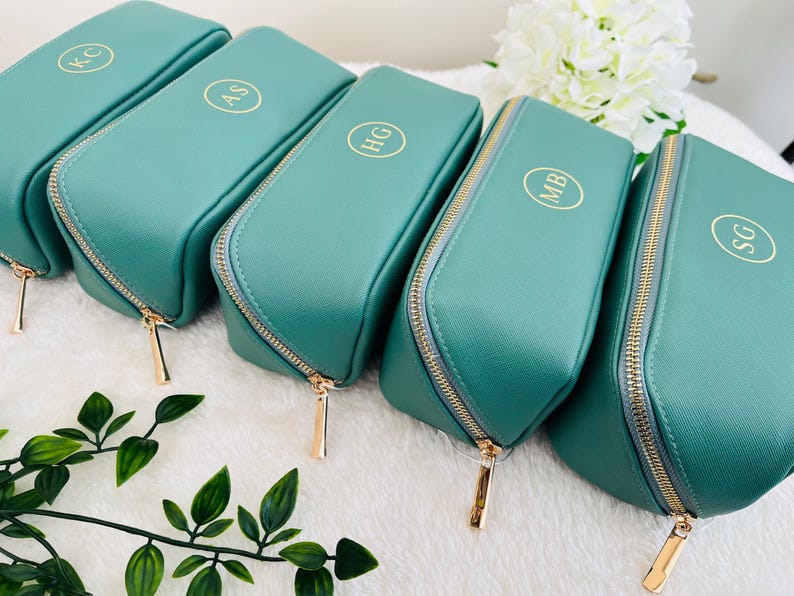 Pu&ograve; includere: Una collezione di trousse per cosmetici color teal con cerniere dorate e iniziali dorate personalizzate. Le trousse sono disposte in fila, mostrando la loro forma rettangolare e la chiusura con cerniera. Le iniziali sono in un cerchio.