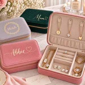 Op de afbeelding: Drie rechthoekige fluwelen sieradendoosjes in roze, grijs en donkergroen, elk met goudkleurige gepersonaliseerde tekst. Een open roze doosje toont gouden kettingen, ringen, oorbellen en een horloge in een beige interieur met vakjes.