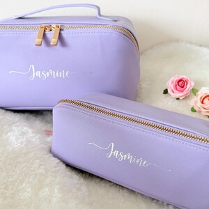May include: Two lavender cosmetic bags with gold zippers and the name "Jasmine" in cursive script. One bag is rectangular with a handle, the other is a smaller, rectangular pencil case. Two pink roses are next to the bags.