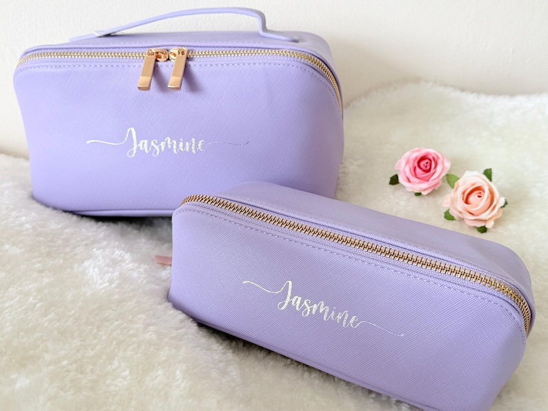 May include: Two lavender cosmetic bags with gold zippers and the name "Jasmine" in cursive script. One bag is rectangular with a handle, the other is a smaller, rectangular pencil case. Two pink roses are next to the bags.
