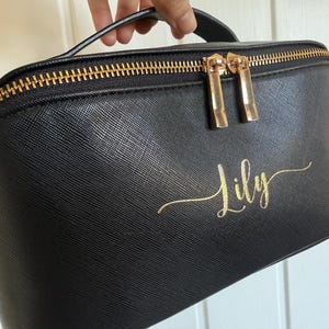 Pu&ograve; includere: Borsa per cosmetici nera con cerniera e manico dorati. La borsa presenta la scritta dorata "Lily". La borsa &egrave; realizzata in un materiale strutturato.
