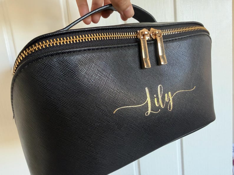 Pu&ograve; includere: Borsa per cosmetici nera con cerniera e manico dorati. La borsa presenta la scritta dorata "Lily". La borsa &egrave; realizzata in un materiale strutturato.