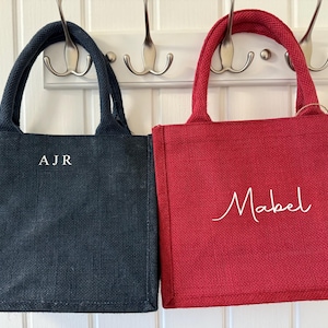 May include: Two small tote bags, one navy blue and one red, hang from hooks. The navy bag has the initials "AJR" in white, while the red bag has the name "Mabel" in white script. Both bags have matching handles.