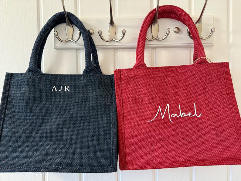 May include: Two small tote bags, one navy blue and one red, hang from hooks. The navy bag has the initials "AJR" in white, while the red bag has the name "Mabel" in white script. Both bags have matching handles.
