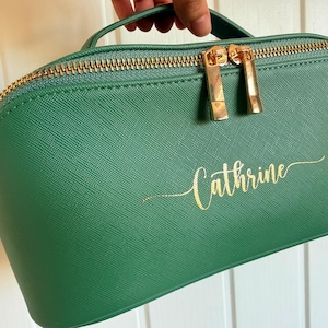Pu&ograve; includere: Un beauty case verde con cerniera e manico dorati. La borsa presenta il nome "Cathrine" in oro sul davanti. I tiranti della cerniera sono dorati.