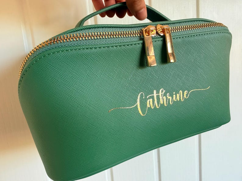 Pu&ograve; includere: Un beauty case verde con cerniera e manico dorati. La borsa presenta il nome "Cathrine" in oro sul davanti. I tiranti della cerniera sono dorati.