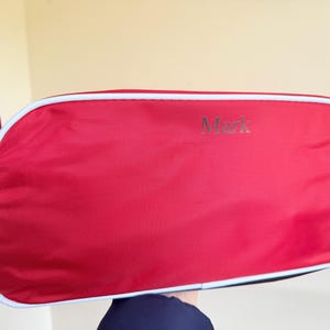 May include: A red rectangular bag with rounded corners and white piping trim. The name Mark is printed in a dark font on the front surface. A black strap is attached to the side, and a red fabric loop extends from the upper left corner.
