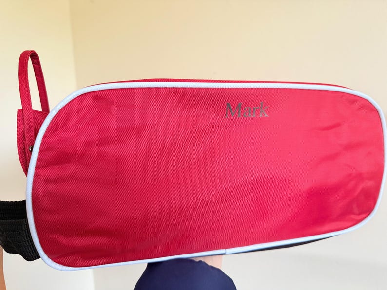 May include: A red rectangular bag with rounded corners and white piping trim. The name Mark is printed in a dark font on the front surface. A black strap is attached to the side, and a red fabric loop extends from the upper left corner.