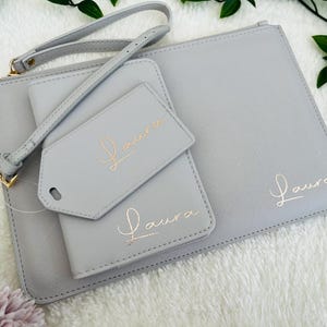 May include: A set of three gray leather travel pouches with gold lettering. The pouches are labeled "Laura" and feature a small tag with the same name.