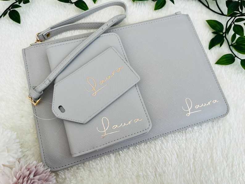 May include: A set of three gray leather travel pouches with gold lettering. The pouches are labeled "Laura" and feature a small tag with the same name.