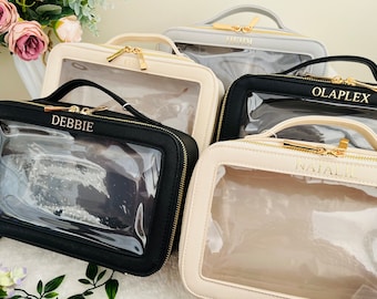 May include: Several cosmetic bags in various colors, including black, cream, and gray, are displayed. Each bag features a clear front panel, a top handle, and a gold-colored zipper. Some bags have names like "DEBBIE" and "OLAPLEX" printed on them.
