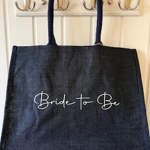 May include: A navy blue tote bag with the words "Bride to Be" in white script. The bag has navy blue handles and is hanging on a white wall with silver hooks.