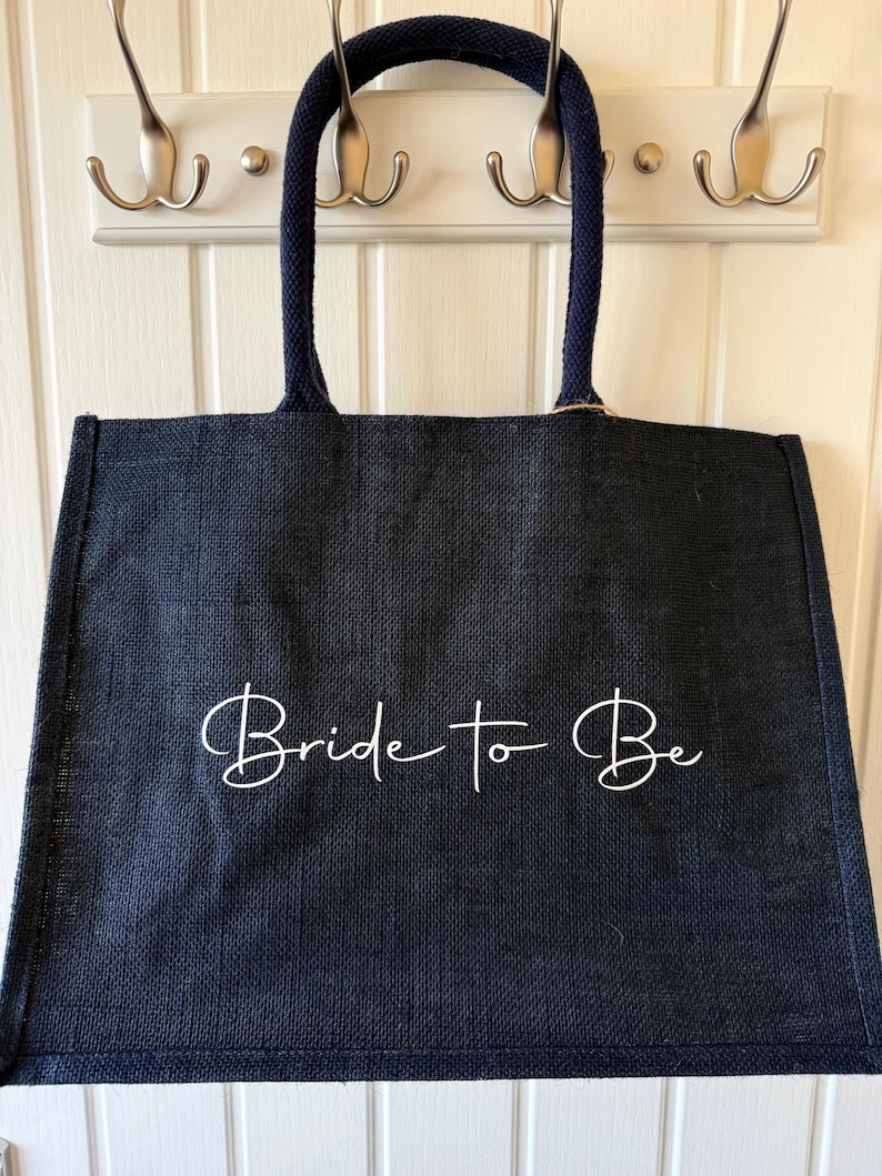 May include: A navy blue tote bag with the words "Bride to Be" in white script. The bag has navy blue handles and is hanging on a white wall with silver hooks.
