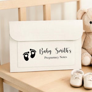 Può includere: Cartella per note di gravidanza color crema con chiusura a patta. La cartella presenta impronte di piedini di bambino nere e il testo "Baby Smith's Pregnancy Notes". Un orsacchiotto e scarpine da bambino sono visibili.