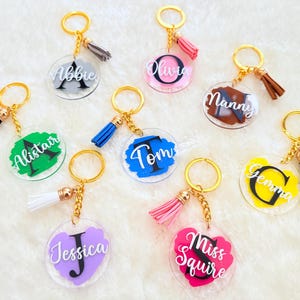 Può includere: Una collezione di portachiavi personalizzati con anelli e nappe color oro. Ogni portachiavi presenta un ciondolo rotondo in acrilico trasparente con un nome o un'iniziale in una varietà di colori, tra cui rosa, blu, verde e giallo.