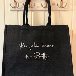 May include: Black tote bag with the text "Le joli bazar de Betty" in white script. The bag has two black handles and is hanging on a white wall with three silver hooks.