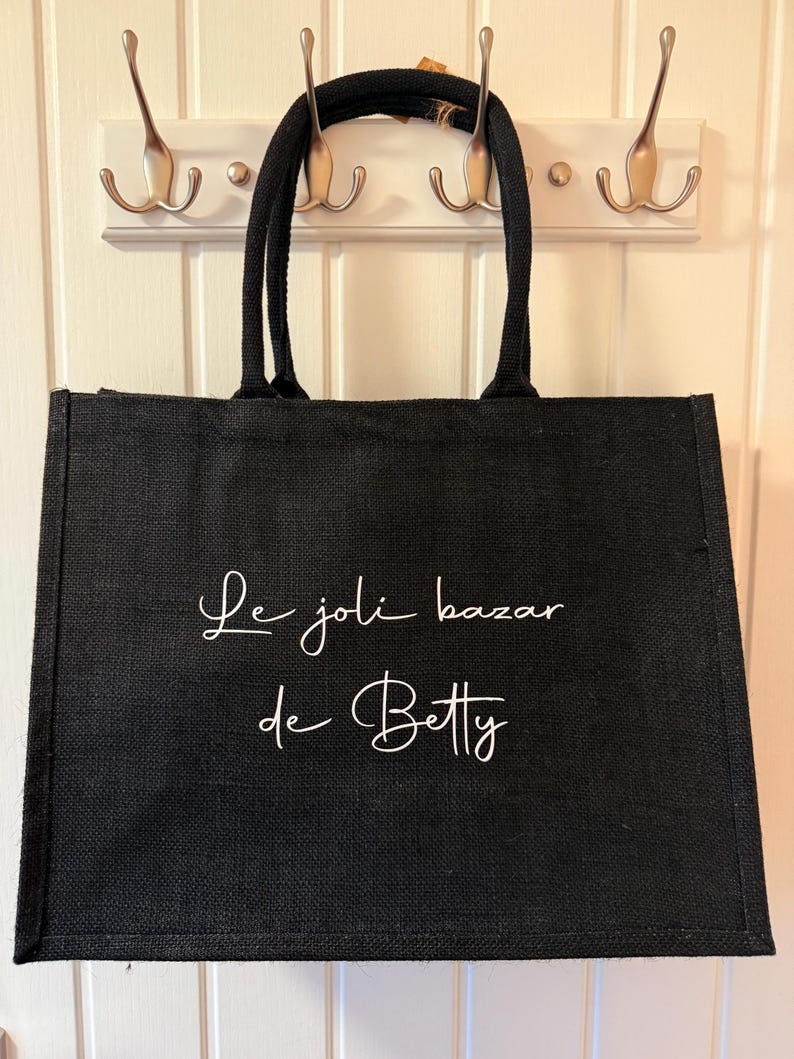May include: Black tote bag with the text "Le joli bazar de Betty" in white script. The bag has two black handles and is hanging on a white wall with three silver hooks.