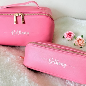 May include: Two pink cosmetic bags with gold zippers and the name "Bethany" in cursive script. One bag is rectangular with a handle, and the other is a smaller, pencil case-style bag. Two pink roses are next to the bags.