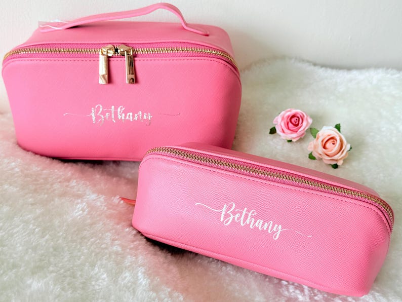 May include: Two pink cosmetic bags with gold zippers and the name "Bethany" in cursive script. One bag is rectangular with a handle, and the other is a smaller, pencil case-style bag. Two pink roses are next to the bags.