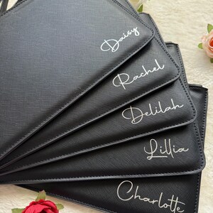 May include: A stack of five black rectangular pouches with a textured finish. Each pouch features a white script name printed on the front: Daisy, Rachel, Delilah, Lillia, and Charlotte. The pouches are arranged in a fan shape.