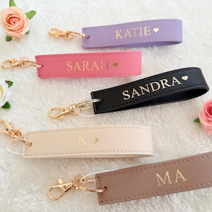 Puede incluir: Cinco llaveros personalizados en varios colores: lavanda, rosa, negro, crema y marrón. Cada llavero presenta un nombre o inicial en letras doradas, con un símbolo de corazón. Cada llavero tiene herrajes dorados.