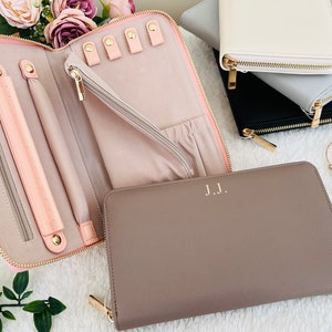 Puede incluir: Un juego de tres estuches para joyas con cremallera en rosa, gris y marrón. El estuche rosa está abierto y muestra un interior de terciopelo rosa con ranuras para anillos y una ranura larga para collares. El estuche marrón tiene las iniciales "J.J." grabadas en dorado en la parte delantera.