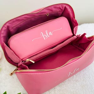 May include: Open pink cosmetic bag with a matching smaller pouch inside. Both pieces have gold zippers and the name "Isla" in white script. The bag is open, revealing the interior lining and compartments.