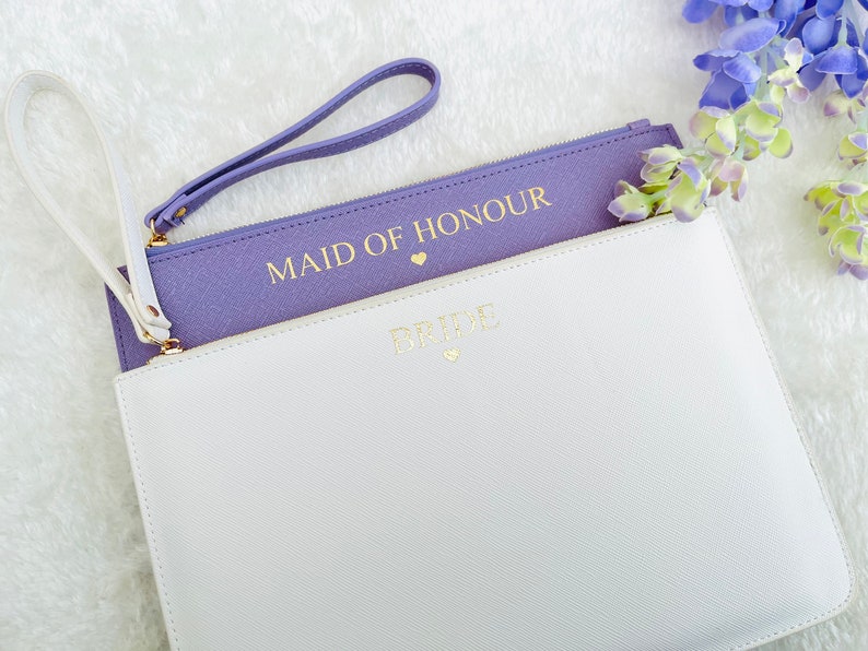May include: Two purple and white zippered pouches with gold lettering. The top pouch says "Maid of Honour" and the bottom pouch says "Bride".