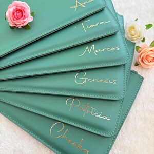 May include: A stack of six sage green rectangular pouches with gold foil lettering featuring the names Ana, Tiara, Marcos, Genesis, Patricia, and Cheska. The pouches are decorated with three artificial roses in pink, white, and peach.