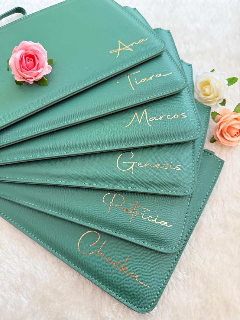 May include: A stack of six sage green rectangular pouches with gold foil lettering featuring the names Ana, Tiara, Marcos, Genesis, Patricia, and Cheska. The pouches are decorated with three artificial roses in pink, white, and peach.