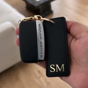 May include: A black Range Rover vehicle key fob with silver accents attached to a rectangular black leather keychain featuring the gold embossed initials SM. Both items are connected by a gold-toned metal ring and clasp hardware.