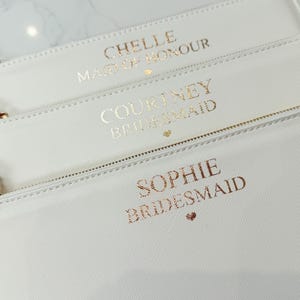 May include: Three rectangular white zippered pouches with wrist straps. Each pouch features metallic gold lettering. The top pouch reads CHELLE MAID OF HONOUR, the middle reads COURTNEY BRIDESMAID, and the bottom reads SOPHIE BRIDESMAID with a small heart.