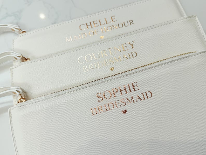 May include: Three rectangular white zippered pouches with wrist straps. Each pouch features metallic gold lettering. The top pouch reads CHELLE MAID OF HONOUR, the middle reads COURTNEY BRIDESMAID, and the bottom reads SOPHIE BRIDESMAID with a small heart.