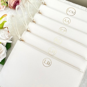 May include: A stack of white rectangular wristlet pouches, each featuring a gold circular monogram with initials including LB, LH, RL, KP, AA, and LCSJ. Each pouch has a matching white wrist strap attached to a gold zipper pull.