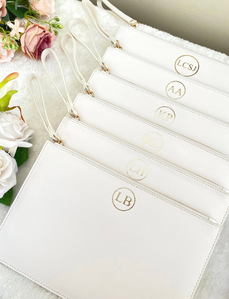 May include: A stack of white rectangular wristlet pouches, each featuring a gold circular monogram with initials including LB, LH, RL, KP, AA, and LCSJ. Each pouch has a matching white wrist strap attached to a gold zipper pull.