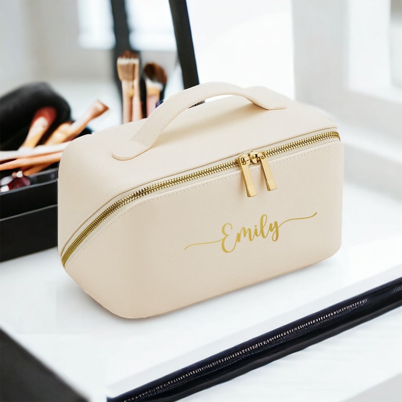 May include: A cream-colored makeup bag with a gold zipper and handle. The bag has the name "Emily" printed in gold script. The bag is rectangular with rounded edges and sits on a white surface.