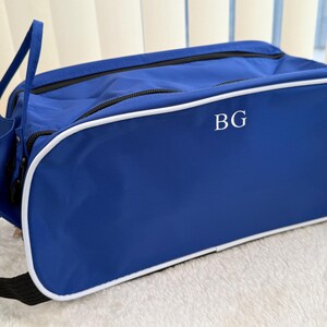 May include: A rectangular blue travel bag with white piping trim along the edges. The front panel features the white embroidered letters BG. The bag includes a black zip closure and a black fabric strap attached to the side for carrying.