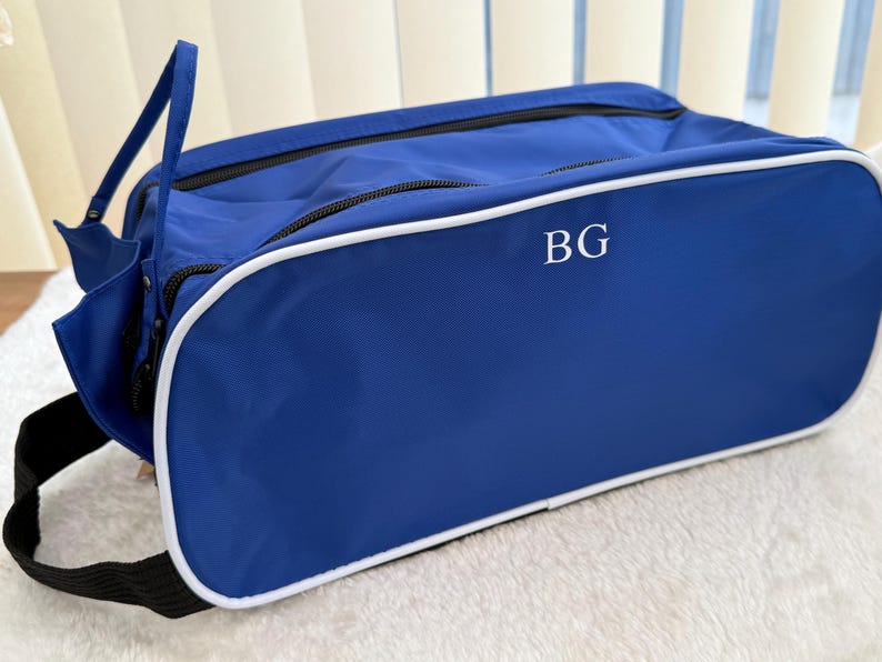 May include: A rectangular blue travel bag with white piping trim along the edges. The front panel features the white embroidered letters BG. The bag includes a black zip closure and a black fabric strap attached to the side for carrying.