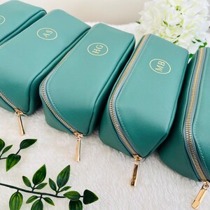 May include: A collection of teal-colored cosmetic bags with gold zippers and personalized initials in gold. The bags are arranged in a row, showcasing their rectangular shape and rounded top. The initials are in a circular design.