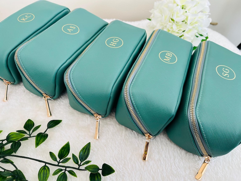 May include: A collection of teal-colored cosmetic bags with gold zippers and personalized initials in gold. The bags are arranged in a row, showcasing their rectangular shape and rounded top. The initials are in a circular design.