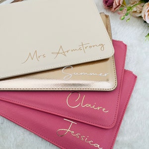 May include: A stack of four rectangular wristlet pouches in cream, metallic gold, and two shades of pink. Each pouch features a personalized name in cursive script: Mrs Armstrong, Summer, Claire, and Jessica, with matching wrist straps.