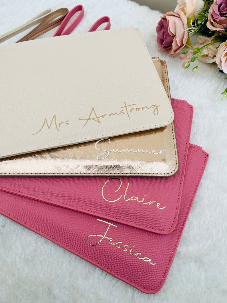 May include: A stack of four rectangular wristlet pouches in cream, metallic gold, and two shades of pink. Each pouch features a personalized name in cursive script: Mrs Armstrong, Summer, Claire, and Jessica, with matching wrist straps.