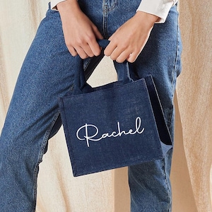 May include: A navy blue tote bag with the name "Rachel" in white script. The bag is held by a person wearing blue jeans and a white long-sleeve shirt. The bag has a rectangular shape and a handle.