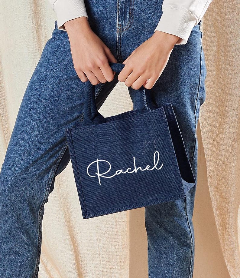 May include: A navy blue tote bag with the name "Rachel" in white script. The bag is held by a person wearing blue jeans and a white long-sleeve shirt. The bag has a rectangular shape and a handle.