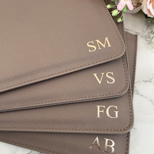 May include: A stack of four taupe-colored rectangular leather-like folders with a cross-hatched texture. Each folder features gold-colored embossed initials in the bottom right corner, reading SM, VS, FG, and AB from top to bottom.