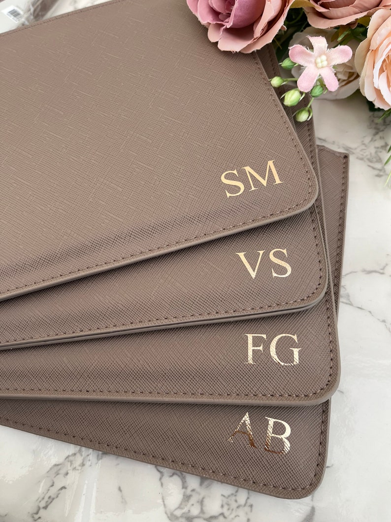 May include: A stack of four taupe-colored rectangular leather-like folders with a cross-hatched texture. Each folder features gold-colored embossed initials in the bottom right corner, reading SM, VS, FG, and AB from top to bottom.