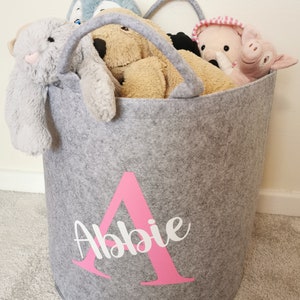 May include: A grey felt storage bin with a pink felt letter A and the name Abbie in white lettering. The bin is filled with stuffed toys, including a grey bunny, a brown dog, a pink pig, and a blue penguin.