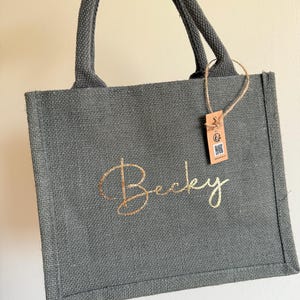 May include: A gray jute tote bag with a gold script name "Becky" on the front. The bag has matching gray handles and a small tag attached. The bag is held up by a hand.